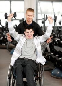 Male NDIS client smiling in a wheelchair holding weights above his head with his disability worker behind him