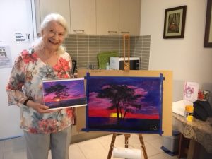 Home Care Package client Lorna Robinson with her painting of a sunset with a tree in the foreground