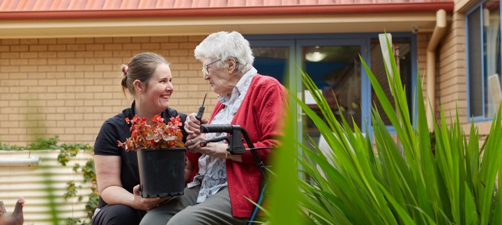 Female home care worker holding a pot plant and kneeling next to female Home Care Package client sitting on a walker